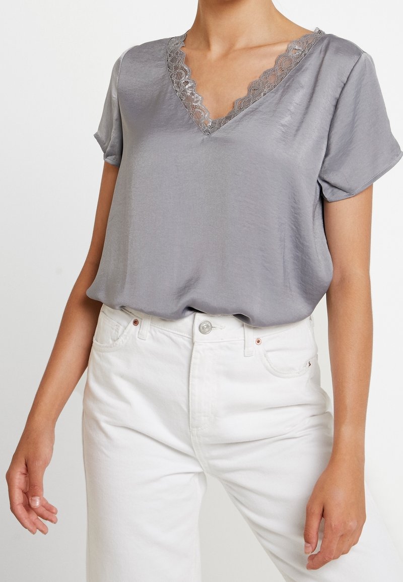 Blusa gris de manga corta con escote en forma de V que presenta detalles de encaje. Textura suave y ajuste relajado. Combinada con pantalones de mezclilla blancos.