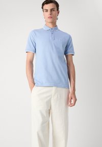 Polo shirt di cotone azzurro chiaro con zip a tre bottoni, colletto a costine e bordi bianchi a contrasto, abbinato a pantaloni color crema.