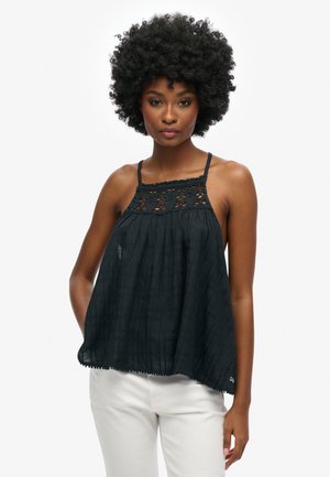 Black sleeveless top with lace detailing at the neckline, pleated fabric, a loose fit, and a scalloped hem. Paired with light-colored pants.