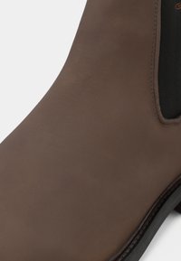 Bottine en cuir marron avec une finition lisse, panneau latéral élastique et accent en textile noir. Semelle brillante avec un léger détail de crantage.