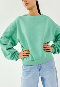 Sudadera verde menta de tejido suave, con un corte relajado, hombros caídos y puños acanalados. Combinada con jeans de color azul claro.