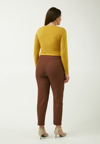 Maglietta a maniche lunghe color senape abbinata a pantaloni marroni aderenti. I pantaloni hanno una texture liscia e l'outfit è completato con scarpe con tacco nude.