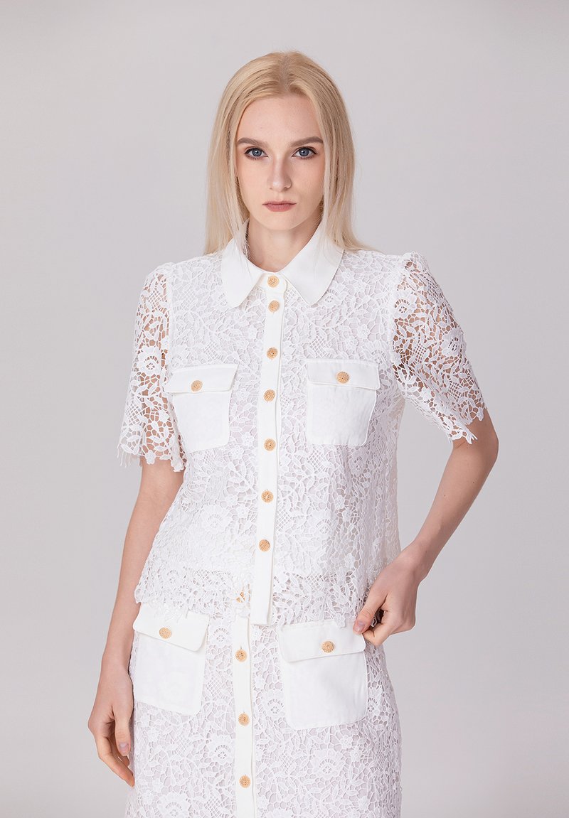 Chemisier blanc en dentelle avec des manches courtes, fermeture à boutons sur le devant, deux poches avant, accents de boutons contrastants, motif floral texturé et col classique.