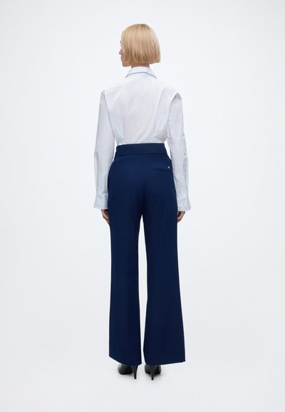Personne aux cheveux blonds courts debout de dos, portant une chemise blanche à manches longues, un pantalon large bleu marine et des talons noirs.
