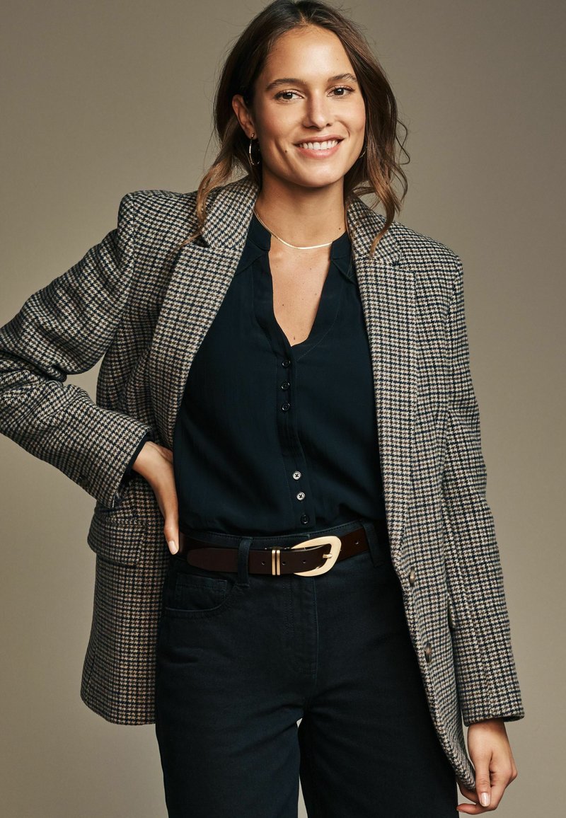 Karierter Blazer in Beige und Braun, über einer schwarzen Bluse. Schwarze High-Waist-Hosen und ein brauer Gürtel mit einer goldenen Schnalle. Glatte Stoffstruktur.