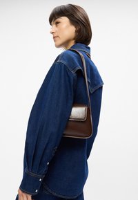 Καφέ δερμάτινη τσάντα με λεία υφή και ορθογώνιο σχήμα, φερόμενη στον ώμο, σε συνδυασμό με ένα γαλάζιο τζιν πουκάμισο με κουμπιά.