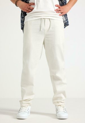 Person iført løse hvide bukser med snøre, hvide sneakers, en hvid t-shirt og en blå mønstret kortærmet skjorte med hænderne på hoften.