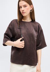 Νεαρή γυναίκα με ίσια μαλλιά, φορώντας γυαλιστερό σκούρο καφέ φαρδύ τοπ, λευκό παντελόνι, ασημένιο βραχιόλι και σκουλαρίκια, κρατώντας το λουράκι μιας μαύρης τσάντας.