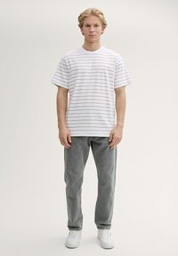 Camiseta blanca con rayas horizontales de color marrón claro, mangas cortas y cuello redondo, combinada con jeans de mezclilla grises y zapatillas blancas.