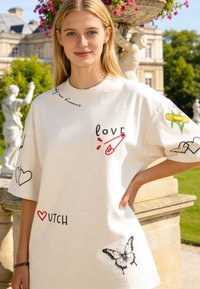 Giovane donna con camicia bianca oversize con amore, farfalla, cuori, bendaggio e testo olandese, in piedi all'aperto vicino a statue e fiori del giardino.