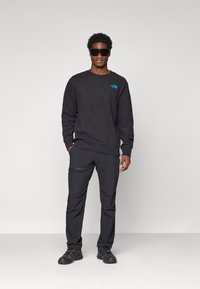 Man in een zwarte sweatshirt met een blauw logo, zwarte broek, zwarte schoenen en grote zwarte zonnebril, staande tegen een effen grijze achtergrond.