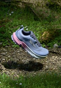 Light gray athletic shoe with pink accents, textured mesh upper, lace-up design, and a cushioned sole, displayed above a grassy background.