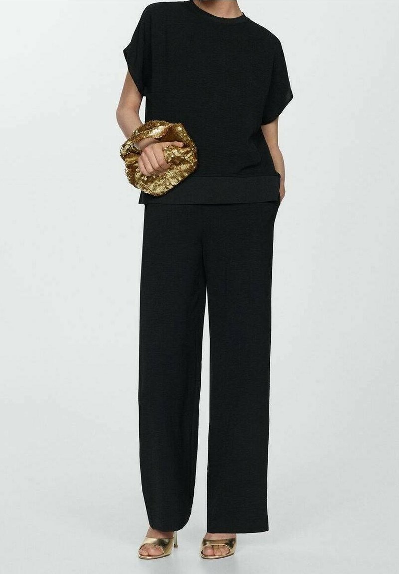 Haut noir à manches courtes avec une coupe ample, associé à un pantalon noir à jambes larges. Une pochette dorée métallique est tenue dans la main gauche.