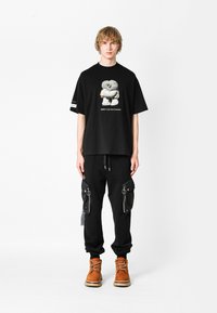 Camiseta gráfica negra con diseño de dibujos animados, combinada con joggers negros con bolsillos con cremallera y botas estilo cordones naranjas. Un atuendo sencillo y casual.