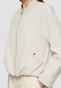 Light beige bomber jacket made of smooth fabric, featuring a full zipper, stand-up collar, and two side pockets with a snap button detail.