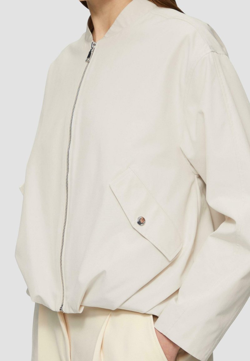 Light beige bomber jacket made of smooth fabric, featuring a full zipper, stand-up collar, and two side pockets with a snap button detail.