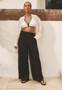 Bílý bikiny top s černým lemováním, spárovaný s volnými černými kalhotami s elastickým pasem. Modelka stojí před světlou zdí.