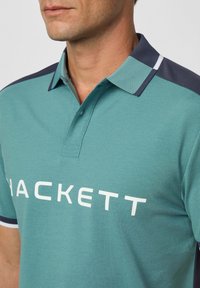 Polo tričko v teal bavlne s námorníckym golierom a bielymi akcentmi. Na prednej strane má veľké biele logo "HACKETT". Štruktúrovaný povrch materiálu.