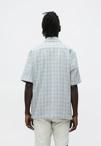 Lühikeste dreadlockidega mees, kes kannab helerohelise triibulise lühikese varrukaga särgi ja beežide lühikeste pükstega, seisab valge taustaga vastu.