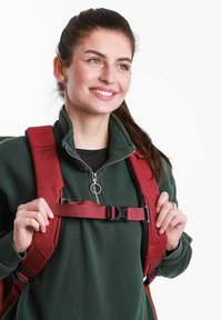 Červený batoh s nastavitelnými popruhy a černými kováními. Model má na sobě zelenou fleecovou bundu, která ukazuje vlastnosti batohu.