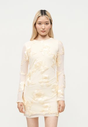 Jeune femme portant une robe blanche transparente à manches longues avec une broderie florale en relief, debout devant un fond blanc uni.