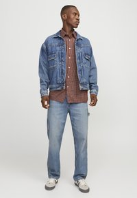 Veste en denim avec poches avant, couleur bleue, et fermeture à boutons, portée par-dessus une chemise à carreaux marron. Associée à un jean de couleur claire et des baskets.