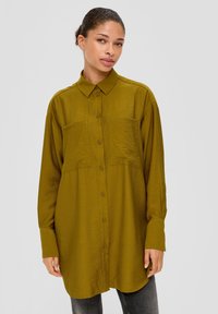 Olijfgroene blouse met lange mouwen, twee borstzakken, knoopsluiting en een ruime pasvorm. De stof lijkt glad met een licht glanzende textuur.