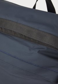Mørkeblå stoffveske med en teksturert overflate, med en horisontal svart stropp og kontrasterende blå sømdetaljer.
