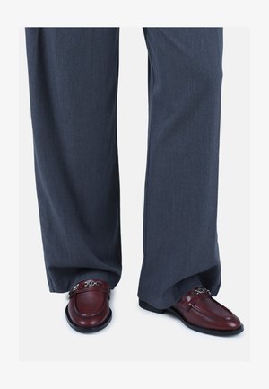 Mocassini in pelle marrone presentano un design semplice con un accento a catena, abbinati a pantaloni a gamba larga grigio scuro realizzati in un tessuto morbido.