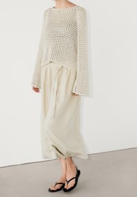 Beżowy sweter z dzianiny z długimi, szerokimi rękawami, noszony z jasnobeżowymi, luźnymi spodniami i czarnymi klapkami, zaprezentowany w minimalistycznym otoczeniu.