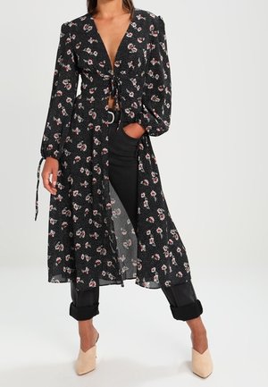 Frau trägt ein langes schwarzes Blumenmusterkleid, das in der Taille über schwarzen Hosen gebunden ist, und beigefarbene spitze Schuhe, steht mit einer Hand in der Tasche.
