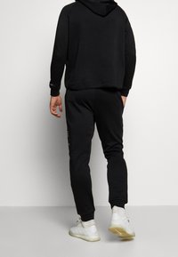 Sweat à capuche noir avec une coupe décontractée et des poignets côtelés, associé à un pantalon de jogging fuselé assorti avec un patch logo latéral et une texture lisse.