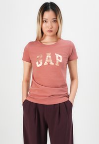 Woman with straight ombre hair wearing a rust-colored GAP t-shirt and dark maroon pants with hands in pockets against white background.