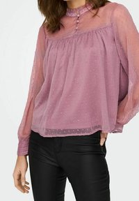 Blouse rose transparente avec pois texturés, col à volants et détail de boutons au niveau de l'encolure ; coupe ample avec manches longues et fluides.