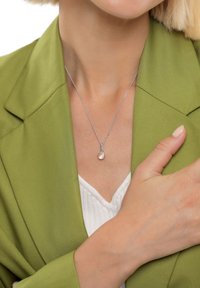 Sudraba kaklarota ar apaļu, gaiši rozā piekariņu, nēsāta virs baltas krekla, papildināta ar zaļu žaketi. Gludas tekstūras un smalka ķēdīte.