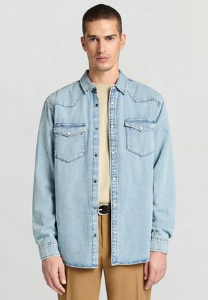 Hellblaues Jeans-Oberhemd mit zwei Brusttaschen, Druckknöpfen und geradem Schnitt. Tragen über einem beigen Rundhalsausschnitt-Top, kombiniert mit hellbraunen Hosen.