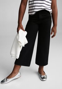Pantalon en denim large noir porté avec un t-shirt rayé. Tenant un pull blanc et portant des ballerines argentées avec un détail en nœud.
