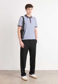 Camisa polo azul-claro com colarinho escuro, textura canelada, calças pretas e ténis brancos. Modelo segura uma pequena mala de ombro preta.