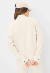 Helle beige Jacke mit strukturiertem Finish, hohem Kragen und Druckknopf-Akzenten an den Ärmelbündchen, getragen mit einer hellbeigen Mütze mit blauen Akzenten.