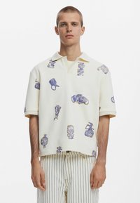 Krém színű póló, lila rajzokkal. Az anyag textúrája érezhető. A modellen csíkos nadrág van. Rövid ujjú és galléros fazon.