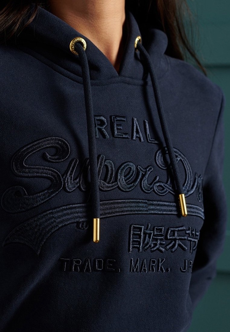 Sweat à capuche bleu marine avec logo brodé "REAL SuperDry", caractères japonais et cordons noirs à embouts dorés autour du cou.