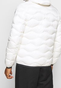 Vit puffjacka med en vågig quiltad design, elastiska ärmslut och en svart fållaccent. Med huva och en slät, glansig yta.