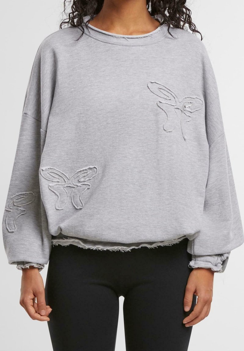 Femme portant un sweat-shirt gris ample avec des découpes en forme de papillon sur la poitrine, la manche et la taille, associée à des leggings noirs.