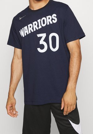 Mann trägt marineblaues Basketballtrikot mit "WARRIORS 30" in Weiß und schwarze Shorts mit weißem Nike-Swoosh am linken Bein.