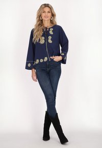 Blusa blu navy con ampie maniche a campana, caratterizzata da ricami dorati e un collo con lacci; abbinata a jeans skinny scuri e stivali neri al ginocchio.