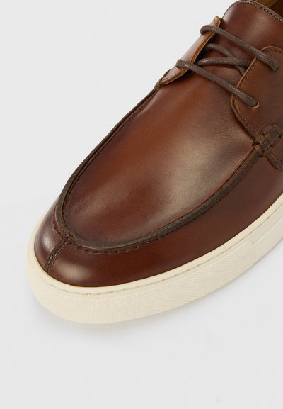 Brown leather shoe with a rounded toe, tonal stitching, and light-colored rubber sole. Features laces and a smooth texture.