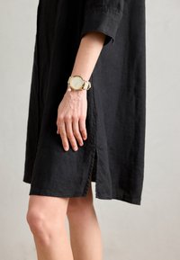 Rochie din in neagră cu mâneci scurte și fente laterale, asortată cu un ceas de mână auriu și crem, cu un cadran rotund și detalii din perlă.