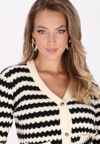Cárdigan a rayas negro y crema con textura acanalada, escote en V y botones de tono dorado. El modelo tiene el cabello ondulado y lleva pendientes de aros dorados.