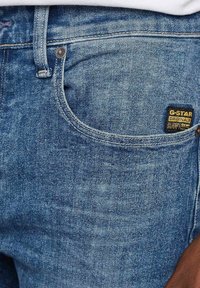 Jeans in denim blu con una texture sbiadita, dotati di una tasca frontale, bottone in metallo e un'etichetta G-Star Originals sulla tasca posteriore.