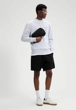 Hombre con gafas, sudadera gris claro, pantalones cortos negros, calcetines blancos, zapatillas blancas, sosteniendo un estuche negro con cremallera sobre un fondo claro y liso.